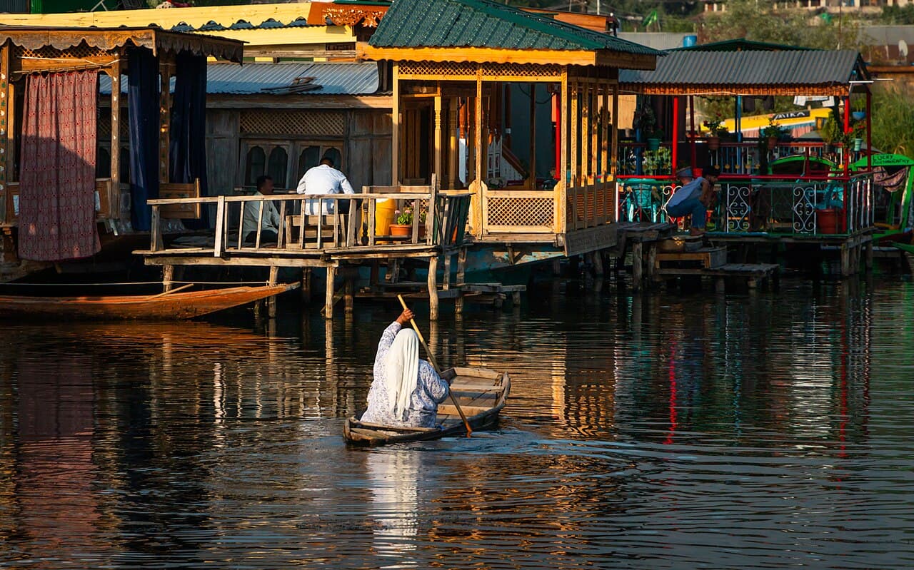 A woman paddles a small boat past wooden houseboats on Dal Lake, Srinagar, in evening light.